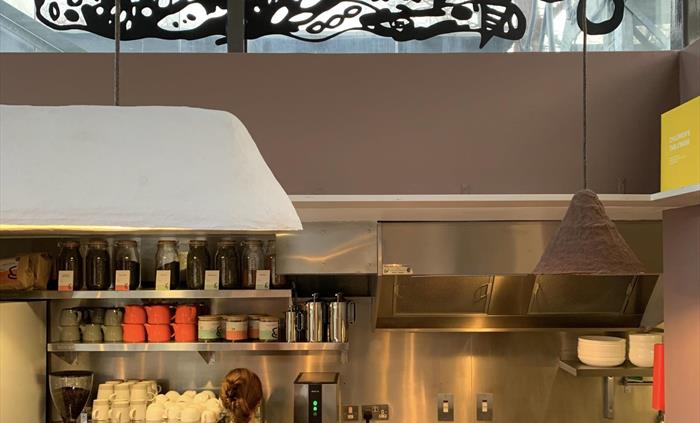 Cafe kitchen with shelves stacked with cups, teapots and jars. a woman with her back to us is operating the coffee machine.